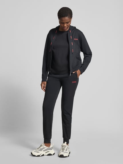 Vrouwenmodel draagt Regular Fit Jogginghose aus Baumwoll-Mix van HUGO, uni zwart, regular fit, mid waist, labelprint, elastische tailleband met trekkoord, zijzakken, sweatstof, katoenmix. Volledig lichaam van voren.