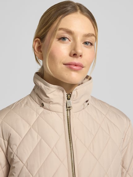 Frauenmodel trägt Steppjacke mit Zweiwege-Reißverschluss von Fuchs Schmitt in Beige, gesteppt, mit Stehkragen, Nahaufnahme Oberkörper. Regular Fit, Kapuze, Eingrifftaschen, Rückenlänge 60 cm, Schulterbreite 41 cm, Brustweite 47 cm, Ärmellänge 63 cm.