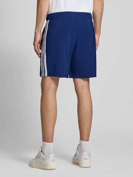 Männermodel trägt Regular Fit Sport-Shorts mit elastischem Bund von adidas Sportswear in Blau mit weißen Kontraststreifen, Ganzkörperansicht Rückansicht.