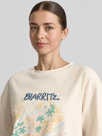 Modelka kobieca nosi damski t-shirt z nadrukiem z motywem i okrągłym dekoltem od Jake*s Casual, bawełna, krój relaxed fit, długość tyłu w rozmiarze S: 58 cm, jasne kolory, prostokątne kształty na nadruku, tekstura gładka. Zbliżenie na górną część ciała.