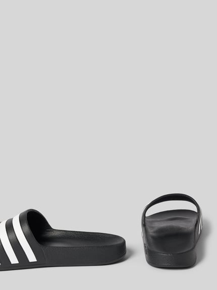 Produktfoto, schwarze Badeschuhe mit weißen Kontraststreifen, abgerundete Schuhspitze, flache Sohle, strukturiertes Material, Adilette Aqua von adidas Sportswear, Damenmodell mit Allover-Muster und offenem Schlupfverschluss.