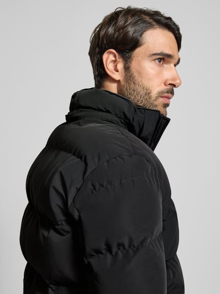 Männermodel trägt schwarzen Mantel mit Kapuze und Label-Patch von NAUTGÅRD, glatte Stoffstruktur, Nahaufnahme Oberkörper, Seitansicht. Gerader Schnitt, Zweiwege-Reißverschluss, seitliche Reißverschlusstaschen, gestrickte Ärmelabschlüsse.