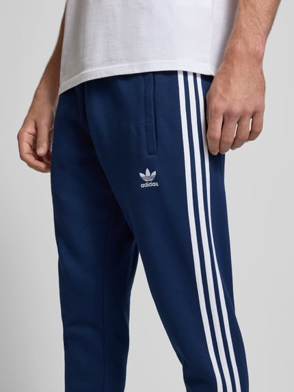 Mężczyzna nosi Spodnie dresowe z elastycznym pasem od adidas Originals, mieszanka bawełny, luźny krój, wpuszczane kieszenie, wyhaftowane logo, długość wewnętrzna nogawki 38 cm, widok z przodu, zbliżenie na dolną część ciała.
