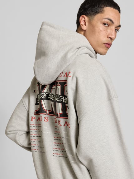 Mannenmodel draagt Oversized Hoodie aus Baumwoll-Mix van RHUDE in lichtgrijs met labelprint, kangoeroezak, ribboorden, ruglengte maat M 64 cm, detailopname rugaanzicht.