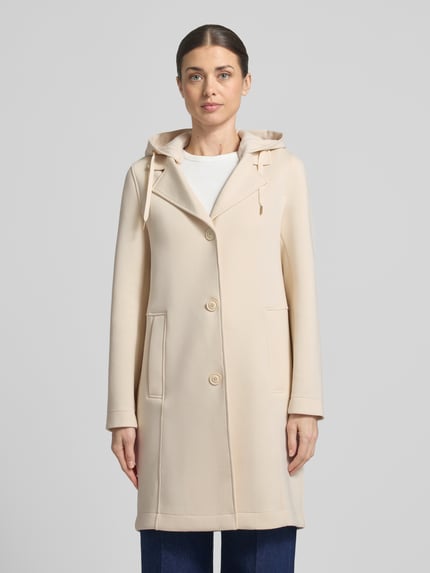 Frauenmodel trägt Regular Fit Kurzmantel mit Kapuze von s.Oliver RED LABEL in Beige mit langen Ärmeln und Leistentaschen, Ganzkörperansicht von vorne.