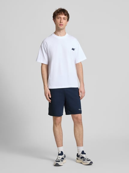 Męski model nosi Shorts mit elastischem Bund und Label-Stitching od CHAMPION, bawełniana mieszanka, krój regular fit, kolor granatowy, szerokość w rozmiarze M: 43 cm, długość nogawki od wewnątrz: 17 cm, widok całej sylwetki z przodu.