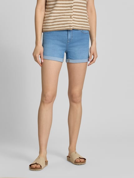 Vrouwenmodel draagt Regular Fit Jeansshorts uit katoenmix model 'CARMEN' van Only, denimlook met labelpatch, 5-pocketmodel, omslag aan de pijpen, knoop- en ritssluiting, vooraanzicht van het hele lichaam.