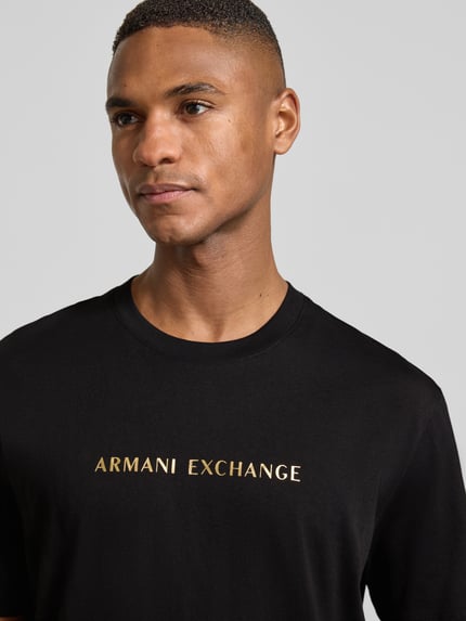 Männermodel trägt T-Shirt mit Label-Print von ARMANI EXCHANGE in Schwarz, reine Baumwolle, Regular-Fit, gerippter Rundhalsausschnitt, Nahaufnahme Oberkörper.