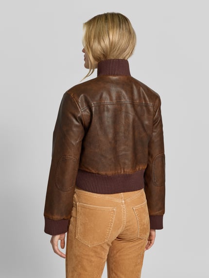 Frauenmodel trägt Lederjacke Modell 'BAILEY' mit Stehkragen von BAUM & PFERDGARTEN in brauner Lederoptik, Struktur, Used Look, mit Ziernähten, Zierknöpfen, metallischen Akzenten, Label-Detail, gerippten Abschlüssen und Taschen, Ganzkörperansicht Rückansicht.