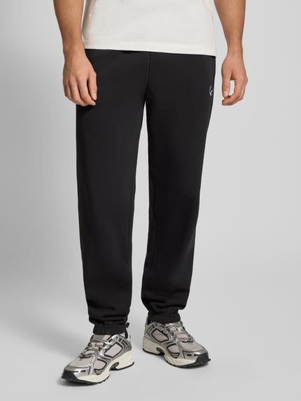 Männermodel trägt Sweatpants mit Label-Stitching von KARL KANI aus Baumwoll-Mix in Schwarz, Regular-Fit, elastischer Bund, Nahaufnahme Unterkörper.
