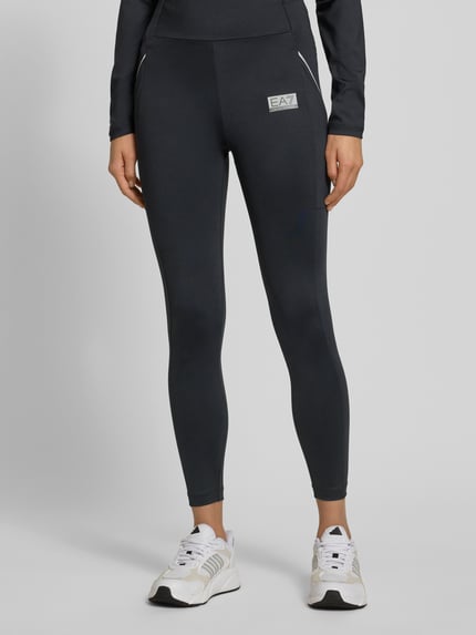 Vrouwenmodel draagt Legging met logo en elastische band van EA7 Emporio Armani, nauwsluitende pasvorm, materiaalmix met stretch, logoprint, elastische band, kleur zwart, textuur glad, detailopname benen, voorkant.