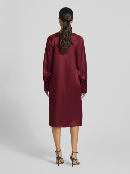 Modelka kobieca nosi Knielanges Cocktailkleid mit Rundhalsausschnitt Modell 'Aurea' od GUIDO MARIA KRETSCHMER WOMAN, bordowy materiał z połyskiem, długi rękaw z mankietem, prosty krój, widoczny materiał-mix ze stretch, zdjęcie całej sylwetki z tyłu.