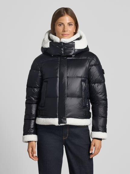 Frauenmodel trägt schwarze Steppjacke mit weißen Fell-Optik-Besätzen, Kapuze und Eingrifftaschen, Steppjacke mit Eingrifftaschen Modell 'NESSIE' von khujo, gerade Passform, Ganzkörperansicht von vorne.