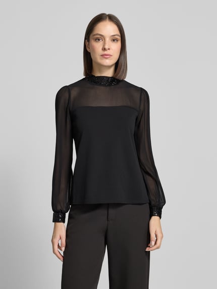 Kobiece modelka nosi bluzkę z długim rękawem z cekinowym obszyciem od Apricot, elastyczna mieszanka materiałów, krój regular fit, stójka, półprzezroczysty materiał na ramionach i bufiaste rękawy, długość tyłu w rozmiarze S: 58 cm, widok całej sylwetki z przodu.