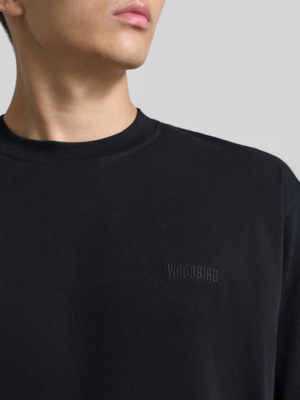 Männermodel trägt Regular Fit T-Shirt aus reiner Baumwolle Modell 'Baine' von WOODBIRD in Schwarz mit Label-Stitching, Nahaufnahme Oberkörper.