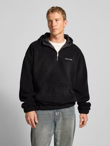 Männermodel trägt Fleecepullover mit Reißverschluss Modell 'Olten' von Pegador in Schwarz mit Kapuze und Känguru-Tasche, Nahaufnahme Oberkörper.