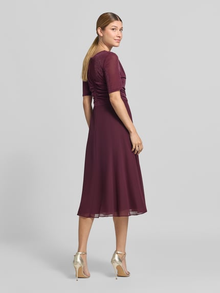 Männermodel trägt Fit and Flare Cocktailkleid mit Wasserfallausschnitt von Vera Mont, sukienka koktajlowa w kolorze bordowym, lekki materiał, gładka faktura, Ganzkörperansicht Rückansicht.