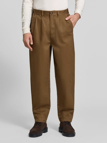 Männermodel trägt Bundfaltenhose mit Twillstruktur von Barbour in Braun, unifarben, Knopfverschluss, Taschen, Ganzkörperansicht von vorne.