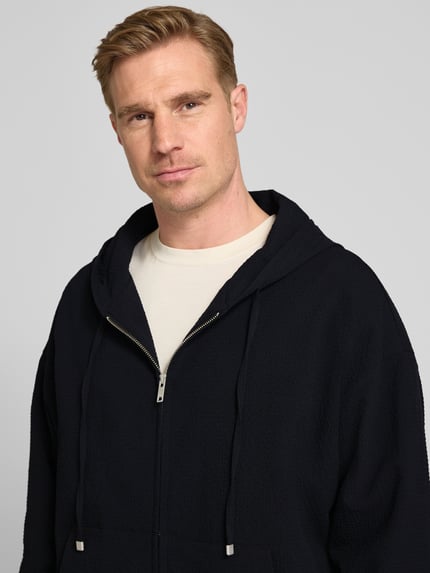 Männermodel trägt Relaxed Fit Sweatjacke aus reiner Baumwolle Modell 'Hootailored262X' von HUGO, efekt: jednolity kolor, zamek błyskawiczny, detal z logo, kaptur z tunel, okrągły dekolt, długi rękaw, zakończenia prążkowane, nakładana boczna kieszeń, dzianina dresowa, bawełna, Nahaufnahme górnej części ciała.