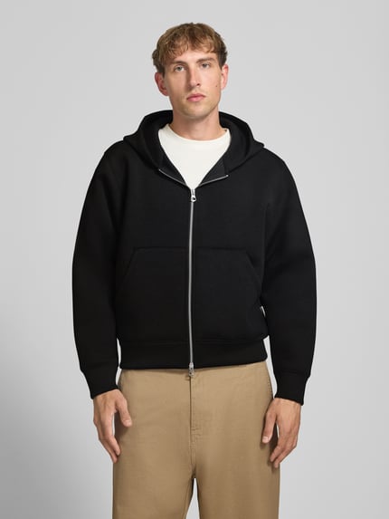Męski model nosi Relaxed Fit Sweatjacke mit Zweiwege-Reißverschluss von JACK & JONES, elastyczna mieszanka materiałów, kieszeń kangurka, zamek błyskawiczny dwustronny, elastyczne wykończenia, Długość tyłu w rozmiarze M: 65 cm, Długość rękawów w rozmiarze M: 65 cm, kolor czarny, gładka tekstura, Ganzkörperansicht von vorne.