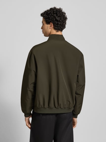 Męski model nosi Regular Fit Jacke mit Stehkragen Modell 'Charge' od Jack & Jones, kolor ciemnozielony, gładka tekstura, widok całej sylwetki z tyłu, krój regular fit, zamek błyskawiczny, kieszenie boczne, elastyczne wykończenia, długość tyłu w rozmiarze M: 66 cm, szerokość ramion w rozmiarze M: 49 cm, szerokość w klatce piersiowej w rozmiarze M: 50 cm, długość rękawa w rozmiarze M: 64 cm.