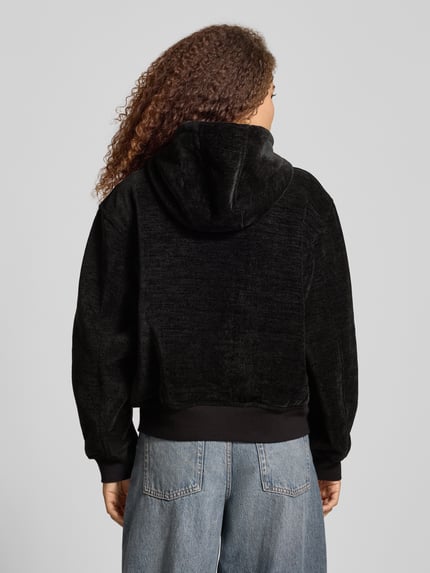 Frauenmodel trägt schwarze Sweatjacke mit Reißverschluss und Känguru-Tasche von Carhartt Work In Progress, Rückenlänge 60 cm, Ganzkörperansicht Rückansicht.