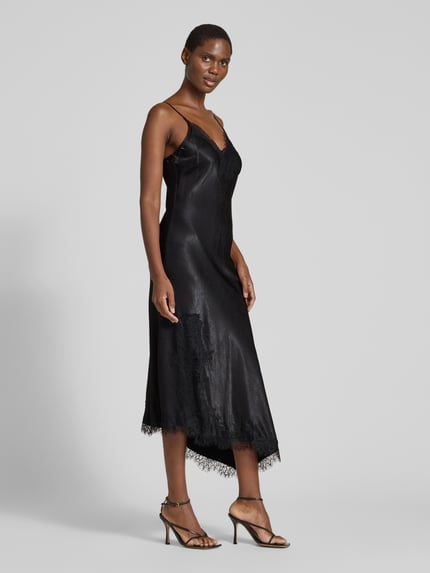 Frauenmodel trägt Maxikleid mit Spitzenbesatz von VERO MODA in Schwarz, glänzende Satinoptik, asymmetrischer Saum, Ganzkörperansicht von vorne.