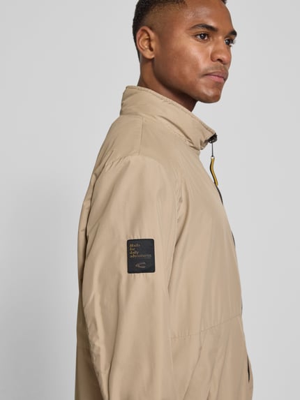 Mężczyznamodel nosi Blouson mit Logo-Print und Stehkragen von camel active, beżowy kolor, gładka faktura, naszywka logo, detal przy zamku, szerokość w rozmiarze 50: 50 cm, długość tyłu: 70 cm, szerokość klatki piersiowej: 58 cm, długość rękawa: 66 cm, zbliżenie na górną część ciała
