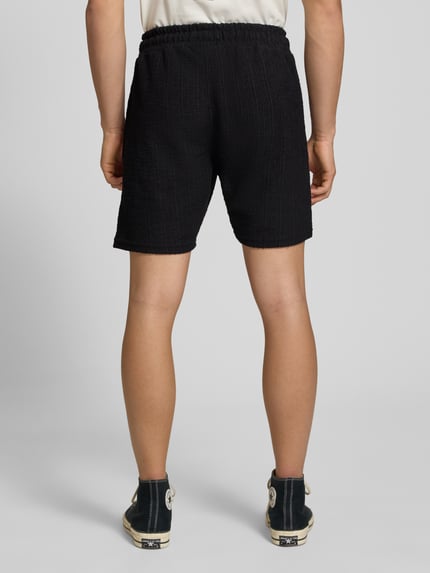 Männermodel trägt Shorts mit Label-Patch Modell 'LIBCO' von Pegador, Schwarz, Strukturmuster, Material-Mix, Gerader-Schnitt, elastischer Bund, Ganzkörperansicht Rückansicht. Kontext: Herren Shorts, Bundweite in Größe M 38 cm, Innenbeinlänge 23 cm.