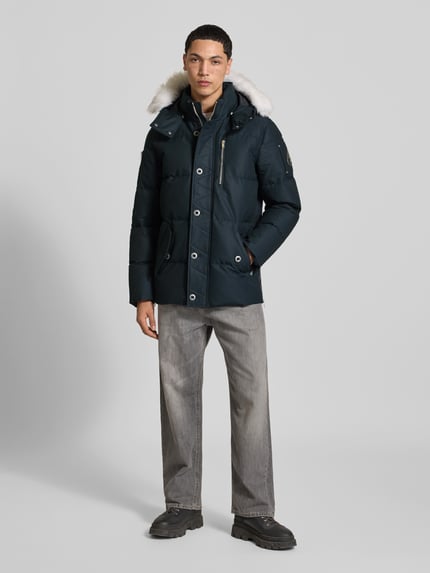 Männermodel trägt dunkelblaue Daunenjacke mit abnehmbarer Fellkapuze von Moose Knuckles, Struktur, Longline, Logo-Patch, Ganzkörperansicht von vorne.