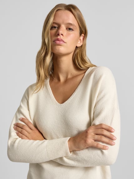 Kobiece modelka ma na sobie Relaxed Fit Pullover mit V-Ausschnitt od ONLY, kremowy kolor, gładka faktura, zbliżenie na górną część ciała.
