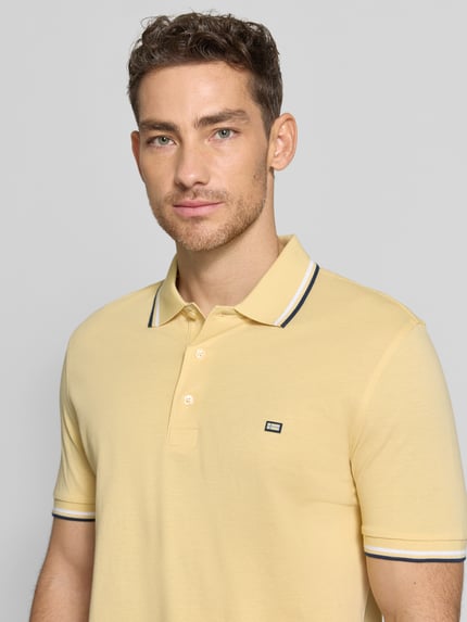 Mężczyznamodel nosi Regular Fit Poloshirt mit Kontraststreifen od Christian Berg Men z bawełny, listwa guzikowa, kolor żółty, długość tyłu 71 cm w rozmiarze M, z przodu na wysokości klatki piersiowej. Nahaufnahme Oberkörper.