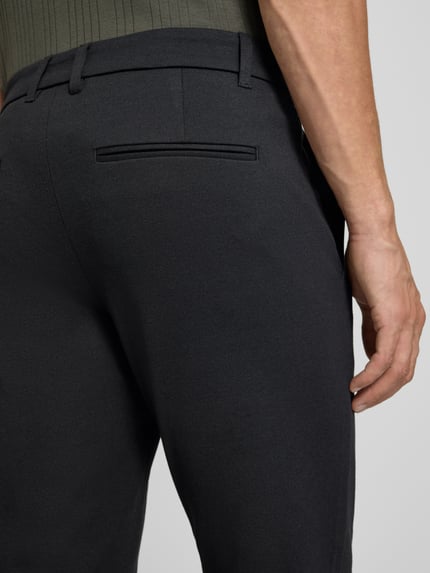 Mężczyznamodel nosi Slim Fit Hose mit Viskose-Anteil in verkürzter Passform von Lindbergh, czarne, gładka tekstura, wpuszczana kieszeń z tyłu, mieszanka wiskozy, krój slim fit, skrócona długość, szerokość w rozmiarze M: 42 cm, długość wewnętrzna nogawki: 72 cm, elastyczna mieszanka materiałów, zapięcie na guzik i zamek błyskawiczny, kieszenie wsuwane, widok z tyłu, detal.