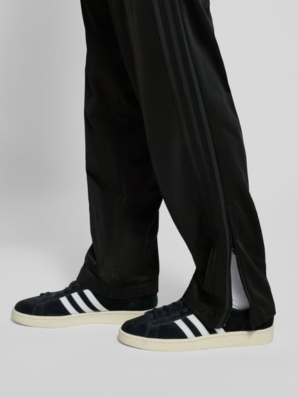 Mężczyznamodel nosi czarne spodnie treningowe z elastycznym pasem adidas Originals, model 'FIREBIRD', regular fit, zbliżenie na nogi.