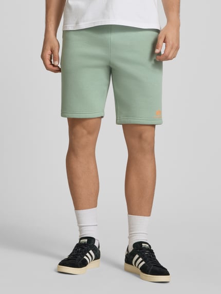 Männermodel trägt Shorts mit Eingrifftaschen und Logo-Print von Alpha Industries in Mintgrün, Relaxed Fit, Ganzkörperansicht von vorne.