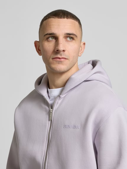 Männermodel trägt Sweatjacke mit Känguru-Tasche von REVIEW in hellem Fliederton, Baumwoll-Mix, Relaxed Fit, Nahaufnahme Oberkörper.
