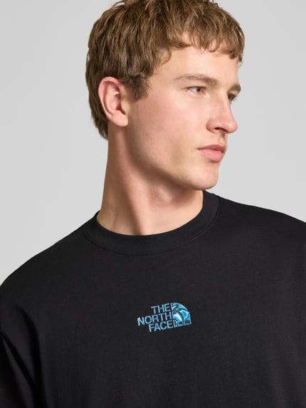 Mężczyznę-model nosi Oversized T-Shirt mit Logo und Rundhalsausschnitt von The North Face, czarna bawełna, logo z przodu, oversize, długość tyłu w rozmiarze M: 73 cm, zbliżenie na górną część ciała