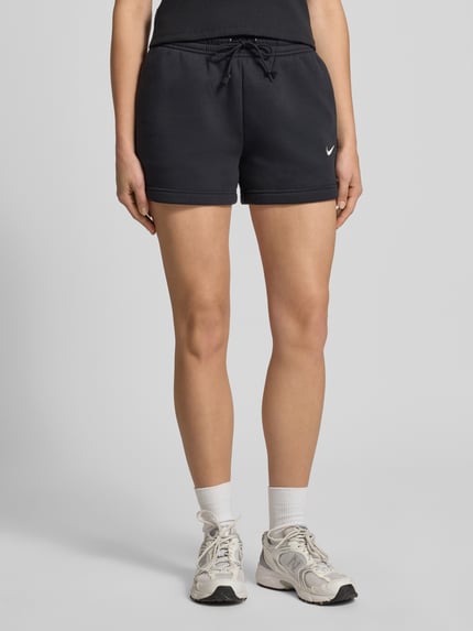 Frauenmodel trägt Sweatshorts mit Logo Modell 'Phoenix' von Nike, Baumwoll-Mix, schwarzer Stoff, Logo links, Ganzkörperansicht von vorne.