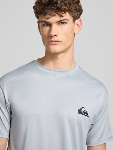 Männermodel trägt T-Shirt mit Rundhalsausschnitt und Logo von Quiksilver, hellgrau, Regular Fit, Material-Mix mit Stretch, Nahaufnahme Oberkörper.