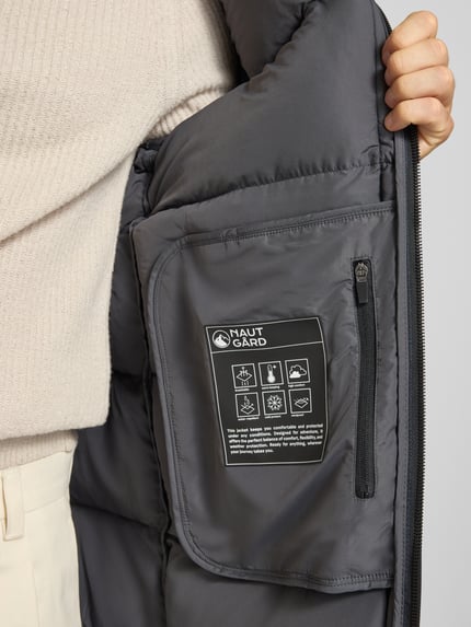 Männermodel trägt Mantel mit Kapuze und Label-Patch von NAUTGÅRD, Farbe Dunkelgrau, glatte strukturierte Oberfläche, Nahaufnahme Oberkörper. Rückenlänge 100 cm, Schulterbreite 52 cm, Brustweite 60 cm, Ärmellänge 65 cm, Zweiwege-Reißverschluss, gestrickte Ärmelabschlüsse, seitliche Reißverschlusstaschen, Label innen.