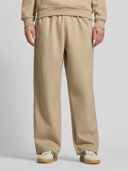 Männermodel trägt Wide Fit Sweatpants mit elastischem Bund von adidas Originals in Beige, Baumwoll-Mix, Logo-Stitching, Nahaufnahme Unterkörper.