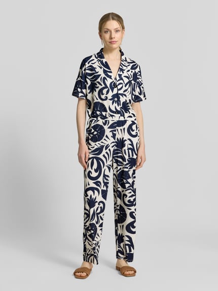 Frauenmodel trägt Jumpsuit mit floralem Allover-Print von Oui, Elastischer-Material-Mix, Taillierte Passform, Reverskragen, Kurzärmelig, Knopfleiste, Farbkombination aus Dunkelblau und Creme, fließende Textur, Ganzkörperansicht von vorne.