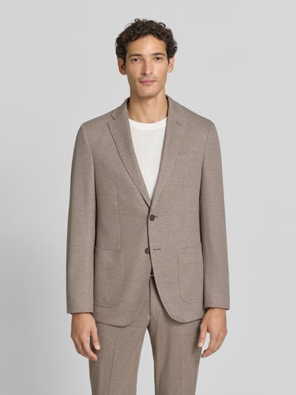 Männermodel trägt fein strukturiertes Slim Fit 2-Knopf-Sakko Modell 'Michel' von Pierre Cardin in Beige, Ganzkörperansicht von vorne. Material-Mix mit Viskose-Anteil, Reverskragen und aufgesetzte Taschen, moderner Look.