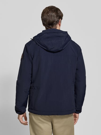 Männermodel trägt Jacke mit Label-Print Modell 'TUNDRA' von Napapijri in Dunkelblau, strukturierte Oberfläche, Ganzkörperansicht Rückansicht. Gerade Passform, Kapuze mit Tunnelzug, Logo-Patch am Ärmel, durchgehender Reißverschluss, Reißverschlusstaschen, elastische Ärmelabschlüsse, Rückenlänge 72 cm, Schulterbreite 48 cm, Brustweite 55 cm, Ärmellänge 63 cm.