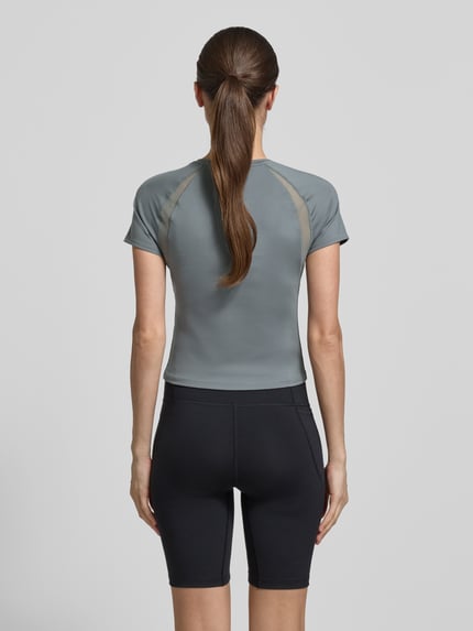Vrouwenmodel draagt T-shirt met Mesh-inzetten en logo van Under Armour, materiaalmix met stretch, kort, ruglengte maat S 45 cm, rond hals, logoprint, mesh-inzetten. Ganzkörperansicht Rückansicht.