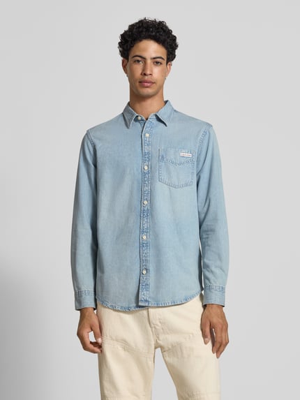 Männermodel trägt Regular Fit Jeanshemd aus reine Baumwolle von Calvin Klein Jeans in denimlook met labelpatch op borstzak, lichte blauwe kleur, lange mouwen met manchet met één knoop, kentkraag, doorlopende knoopsluiting, materiaal puur katoen, ganzkörperansicht von vorne.
