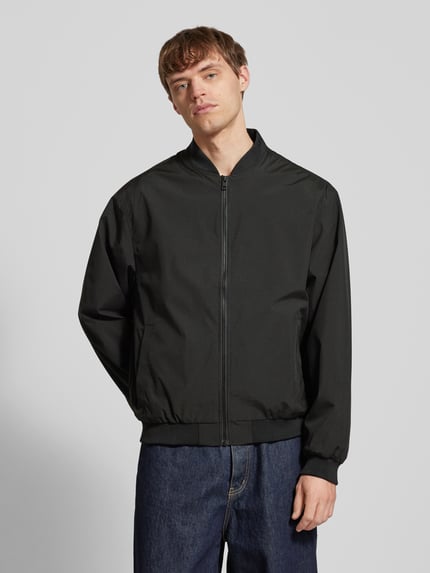 Męski model nosi prosty bomber CHARGE marki Jack & Jones, kolor czarny, kieszenie boczne, ściągacze, długość tyłu 66 cm, szerokość ramion 54 cm, szerokość klatki 65 cm, długość rękawa 64 cm, krój straight fit, zamek, widok całej sylwetki z przodu.
