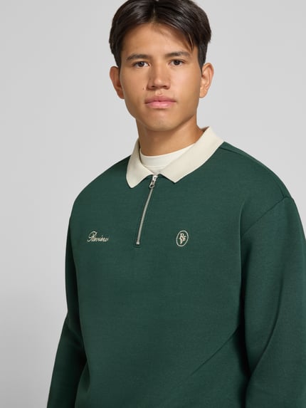 Männermodel trägt Regular Fit Sweatshirt aus Baumwoll-Mix von REVIEW in Dunkelgrün mit Polokragen, Logo-Stitching, Nahaufnahme Oberkörper.