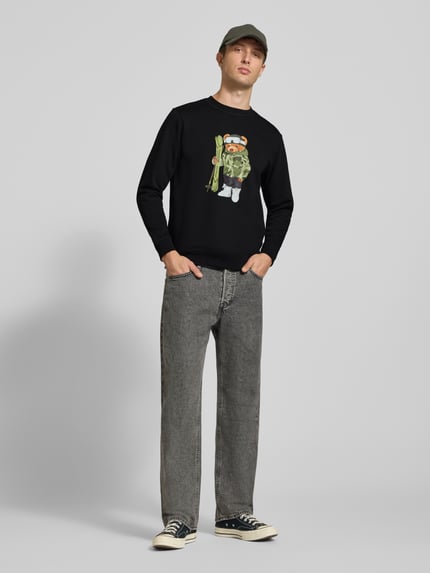 Męski model nosi bluzę Sweatshirt mit Motiv-Print od CHILL PILL, mieszanka bawełny, krój regular fit, okrągły dekolt, długość tyłu w rozmiarze M: 68 cm, długość rękawa: 66 cm, czarny kolor z nadrukiem z motywem, pełny widok z przodu.