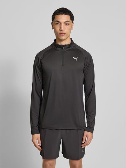 Męskamodel nosi bluzkę z długim rękawem 'Langarmshirt mit Stehkragen und Reißverschluss' od PUMA PERFORMANCE, krój regular fit, stójka, zamek błyskawiczny, logo, długość tyłu w rozmiarze M: 71 cm, widok całej sylwetki z przodu.